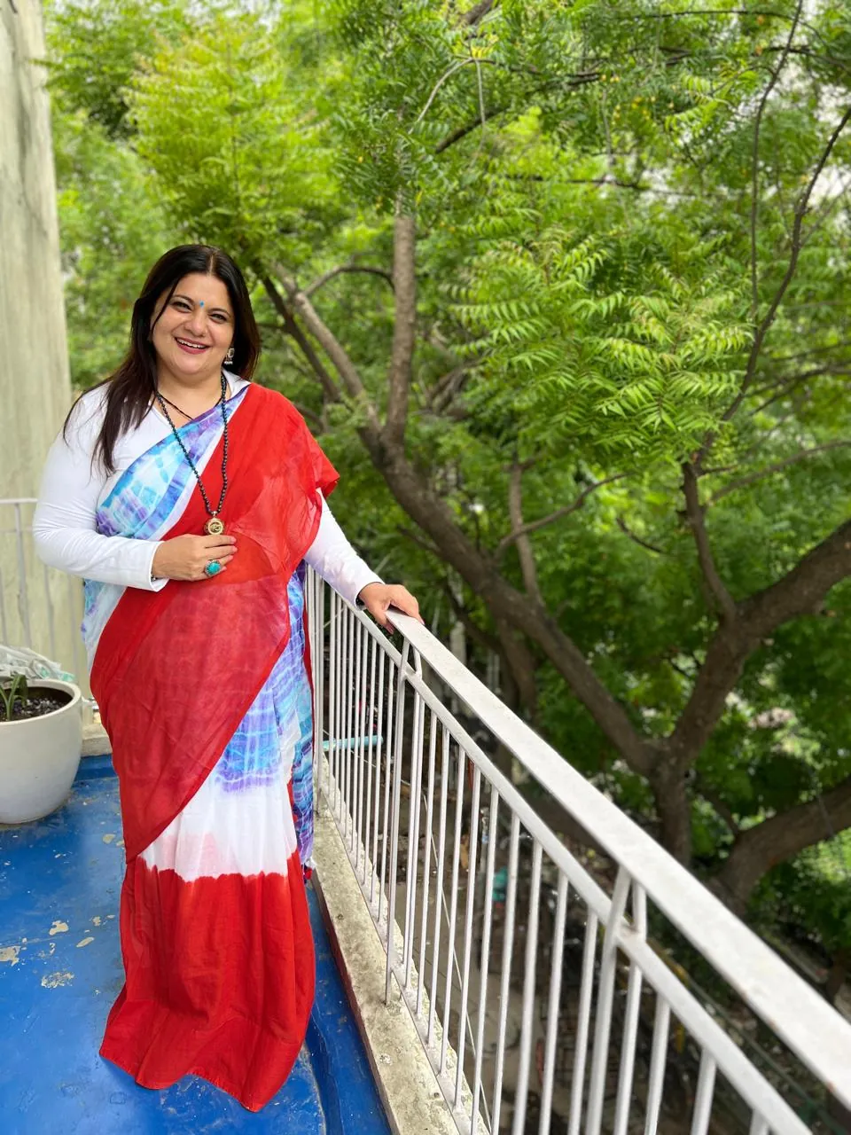 Sapnaa Khemani on a balcony in a red and blue tie-dye saree, trees behind her